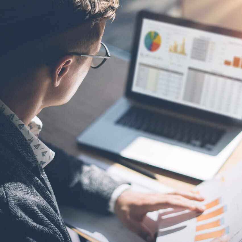 Young finance market analyst in eyeglasses working at sunny office on laptop while sitting at wooden table.Businessman analyze document in his hands.Graphs and diagramm on notebook screen.Vertical